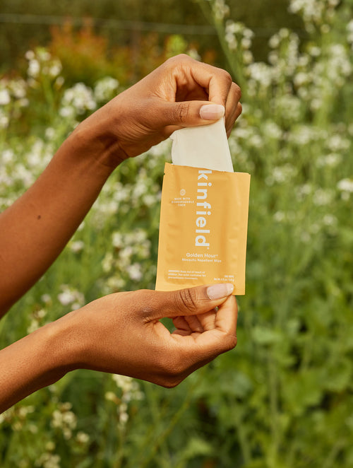 A women's hands open an orange Golden Hour Wipe packet and pulls out the biodegradable repellent wipe in front of greenery.