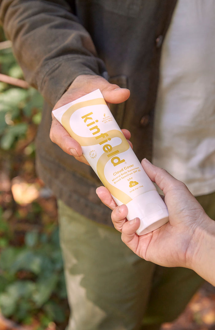 A hand reaches for a tube of Kinfield Cloud Cover Body Sunscreen being handed to her by another hand.