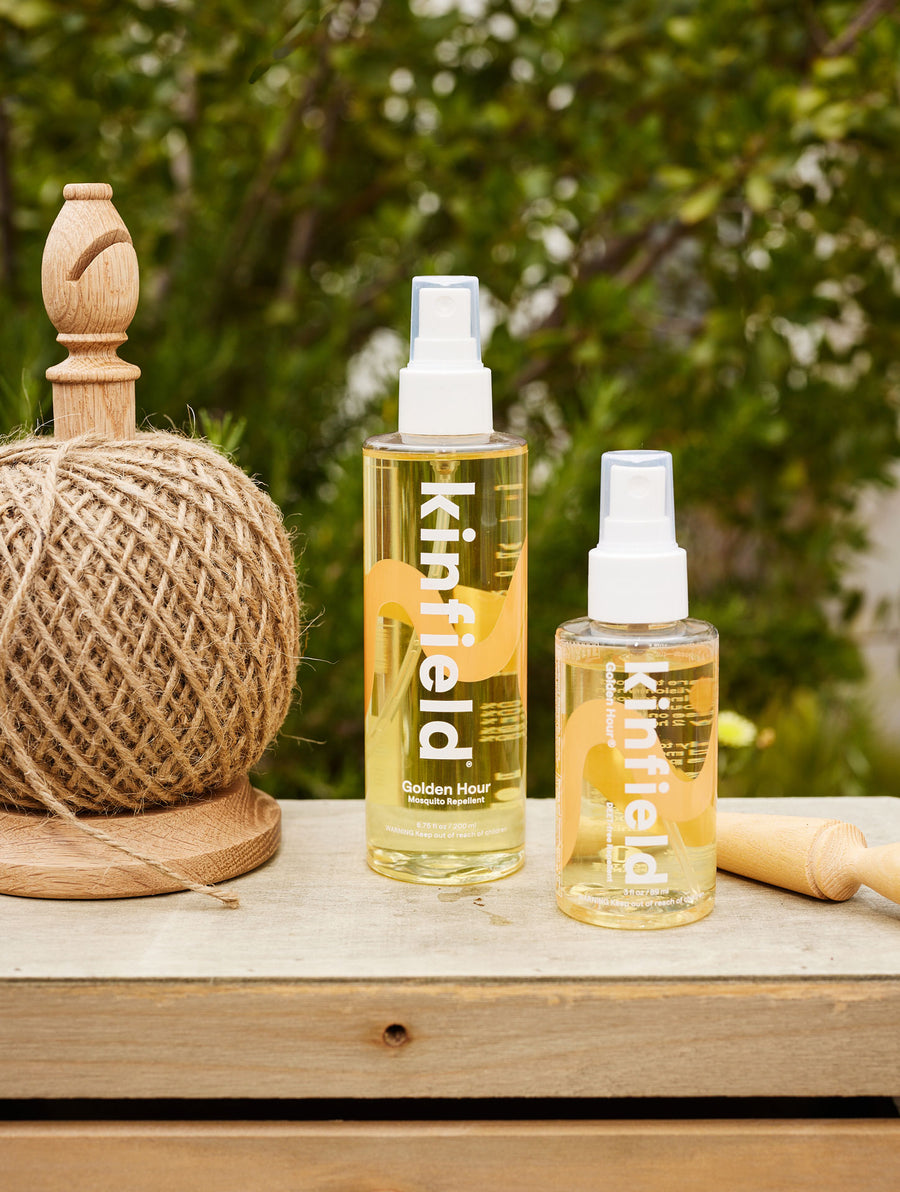 Two bottles (one 3 oz, one 6.75 oz) of Kinfield Golden Hour sit on a wood box in front of a greenery backdrop next to twine.