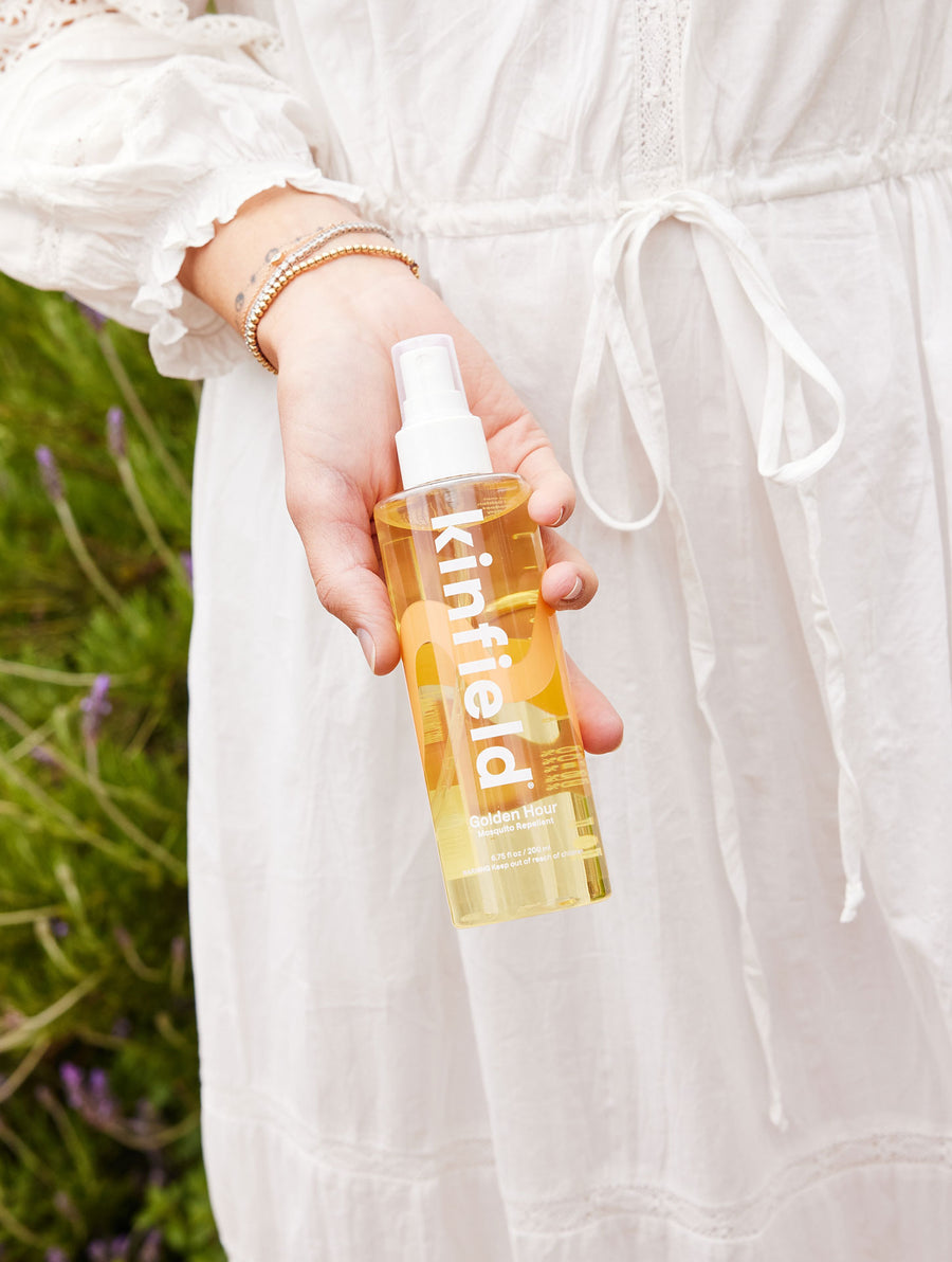 A woman's hand holds out a 6.75 oz bottle of Kinfield Golden Hour while wearing a white dress in front of greenery.