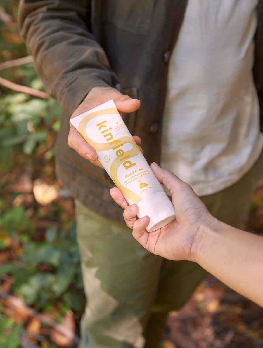 Two people pass a tube of Cloud Cover Sunscreen between them in the woods.