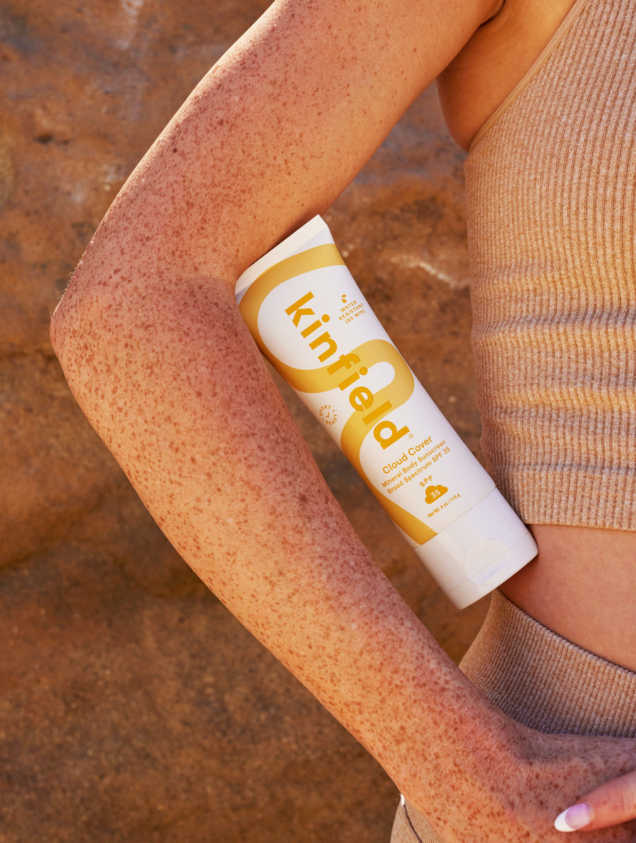 Cloud Cover Sunscreen rests in the crook of a woman's freckled elbow in front of a rock on a sunny day.