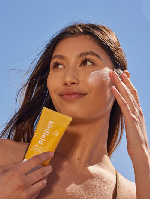 A woman holds a tube of Kinfield Daily Dew SPF 35 against a blue sky, with a sheer swish of product on her cheek.