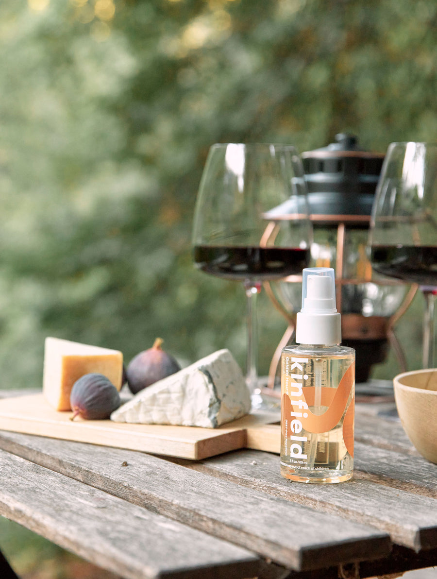 A bottle of Kinfield Golden Hour rests on a wooden table next to a cheese plate and two glasses of wine.