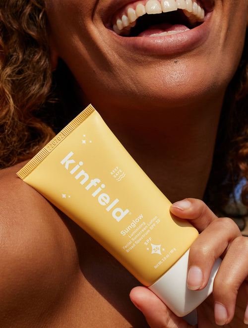 Close up photo of Kinfield Sunglow SPF 35 held up by a smiling woman with curly hair