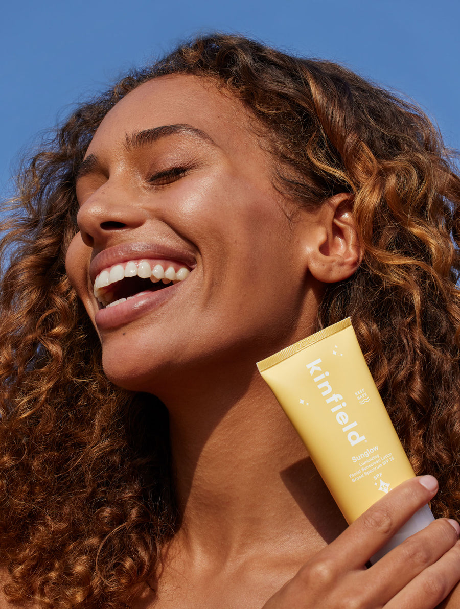 Smiling woman with curly hair holds up Kinfield Sunglow SPF 35