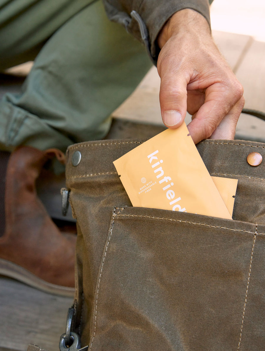 Man's hand pulls a Kinfield Golden Hour Repellent Wipe from his brown textured bag.