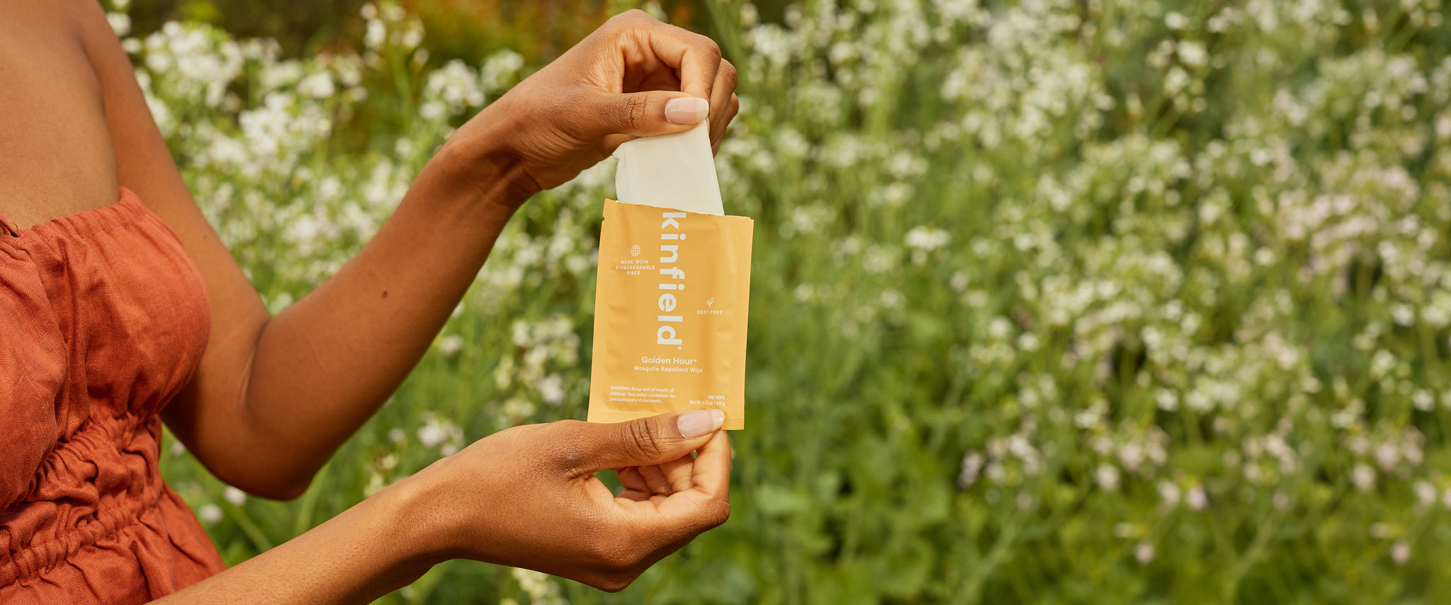 A women's hands open an orange Golden Hour Wipe packet and pulls out the biodegradable repellent wipe in front of greenery.