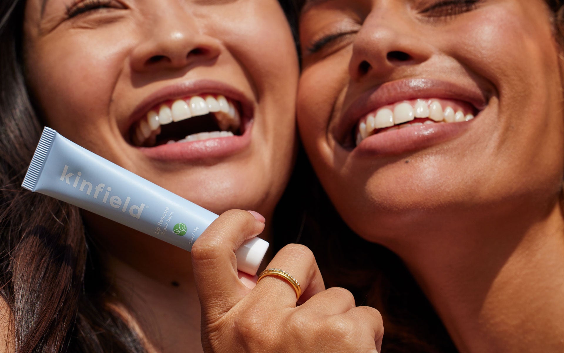 Two women are smiling brightly; one holds Kinfield Lip Rescue near her face.