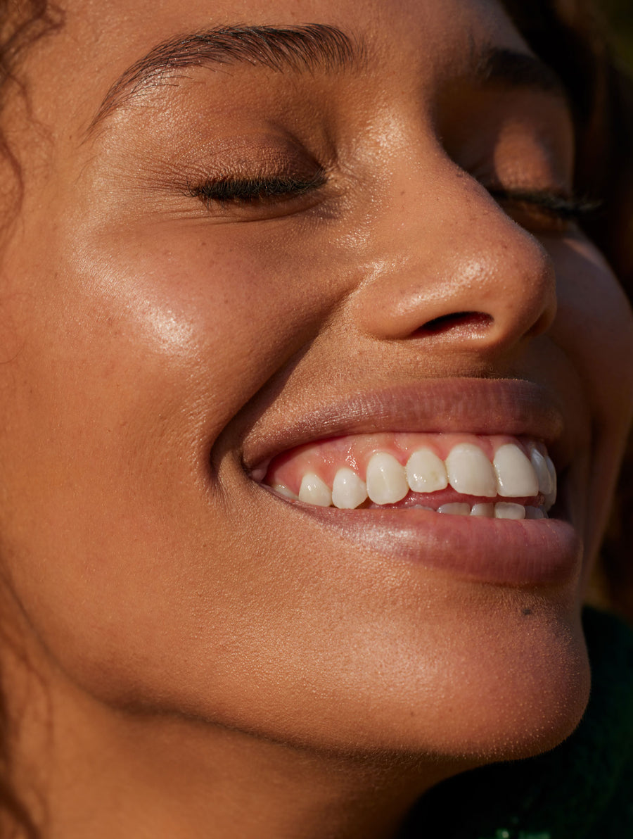 A woman with glowing brown skin smiles brightly into the sun.